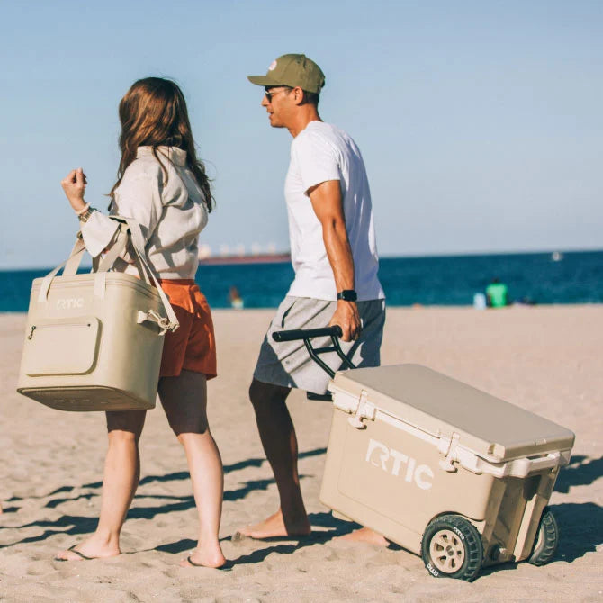 Two people walk on a sandy beach carrying a cooler bag and a wheeled cooler, with the ocean visible in the background.